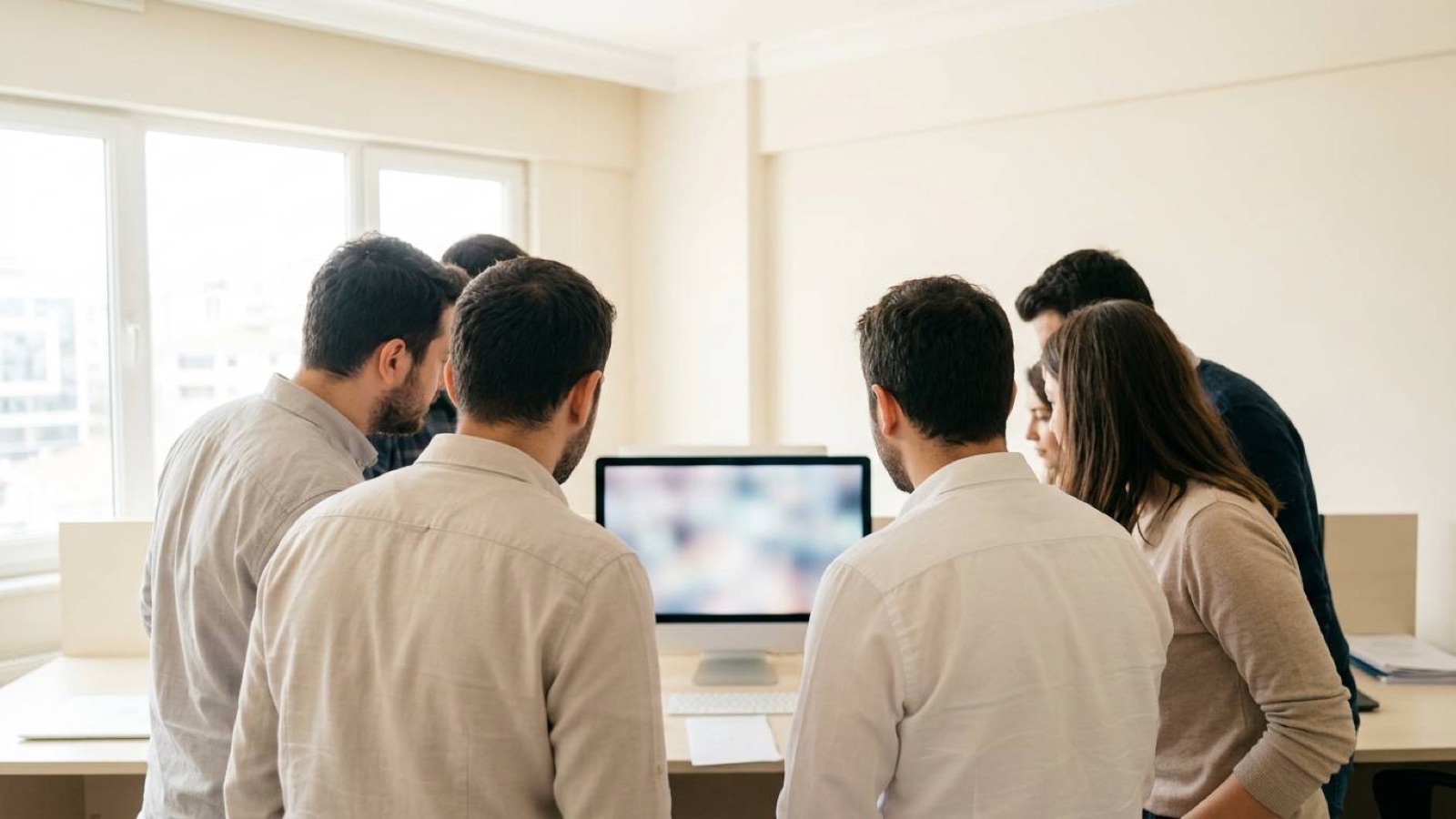 Six colleagues gathered around a single monitor in a bright office, seen from behind