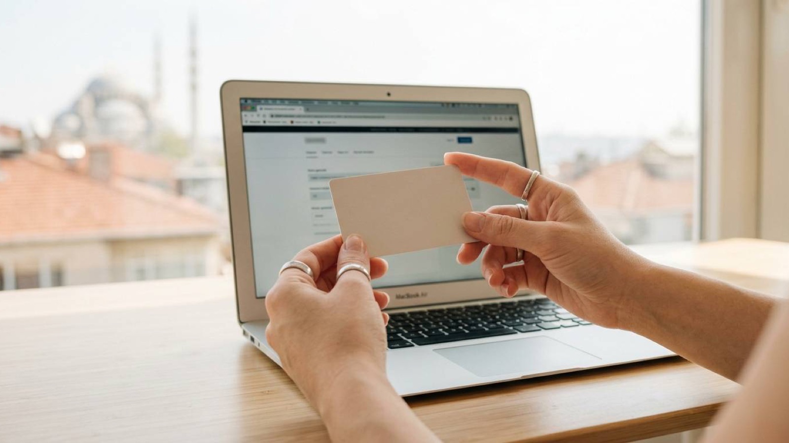 Hands holding a blank payment card in front of an open laptop, Istanbul rooftops through the window