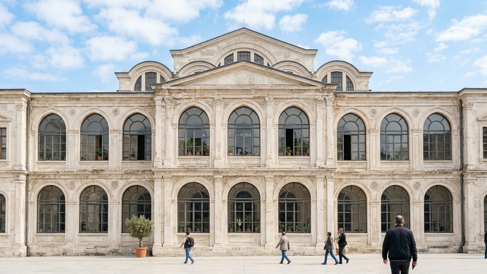 The stone facade of a nineteenth century public building with tall arched windows, people crossing the plaza in front