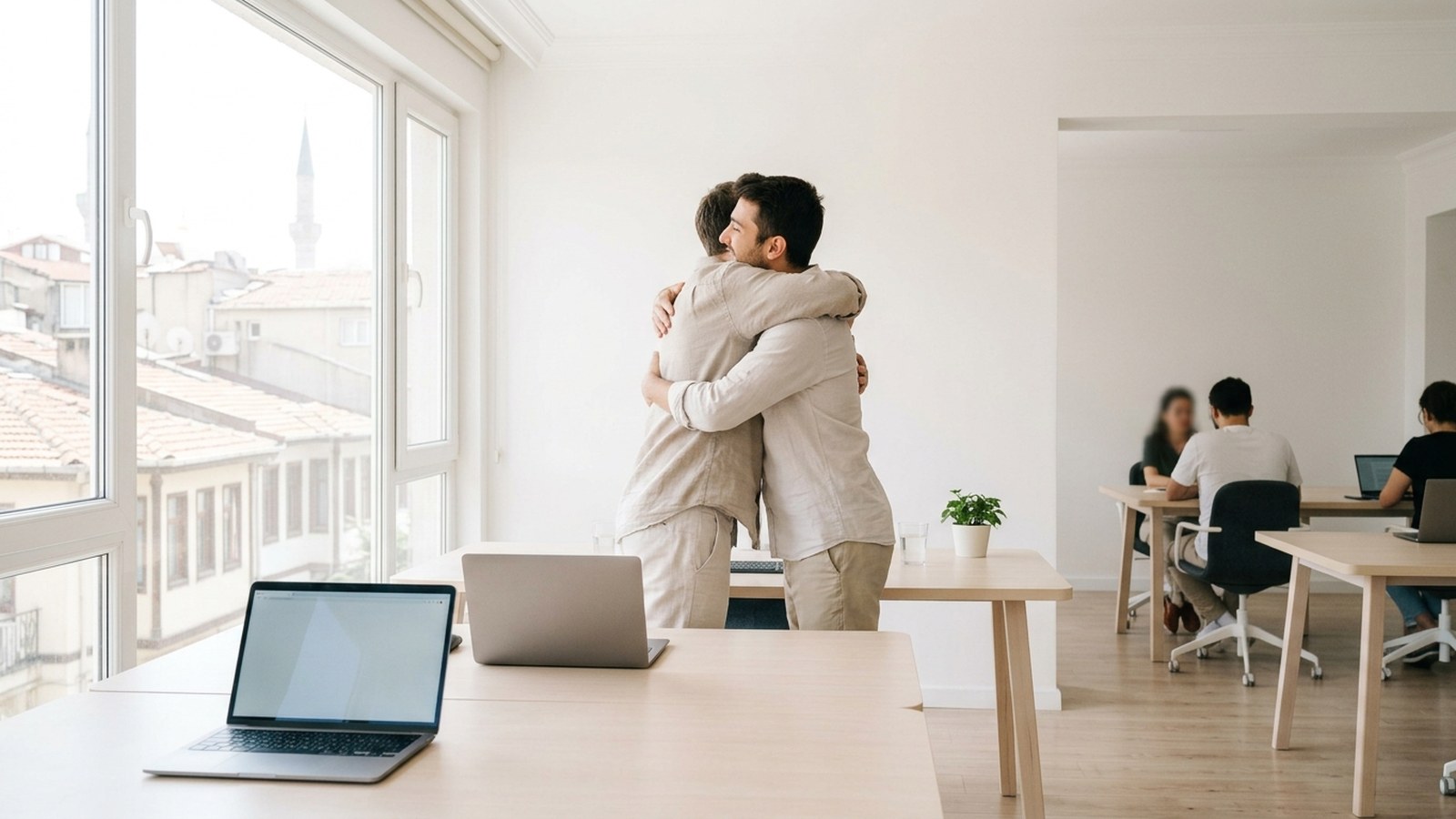 Two colleagues embracing in a bright office with laptops open on the table in front of them