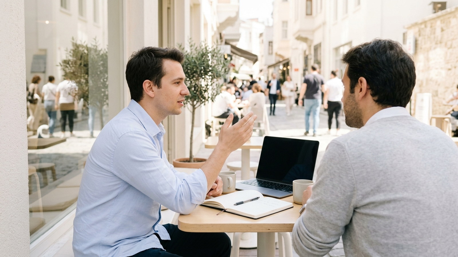 Two men talking across a small round table on a sunlit street terrace, one gesturing mid-sentence, a laptop and notebook between them